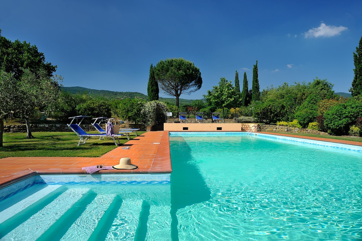 The outdoor pool area is surrounded by lush greenery, featuring comfortable lounge chairs and umbrellas for relaxation. Steps lead gently into the clear blue water, while a sunhat rests on the pool’s edge, inviting guests to enjoy the serene atmosphere.