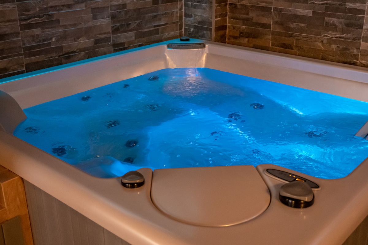 A modern hot tub is situated in a dimly lit space, featuring illuminated water in calming blue tones. The intricate tile backdrop complements the sleek design, creating a relaxing atmosphere around the tub, which is equipped with control buttons for ease of use.