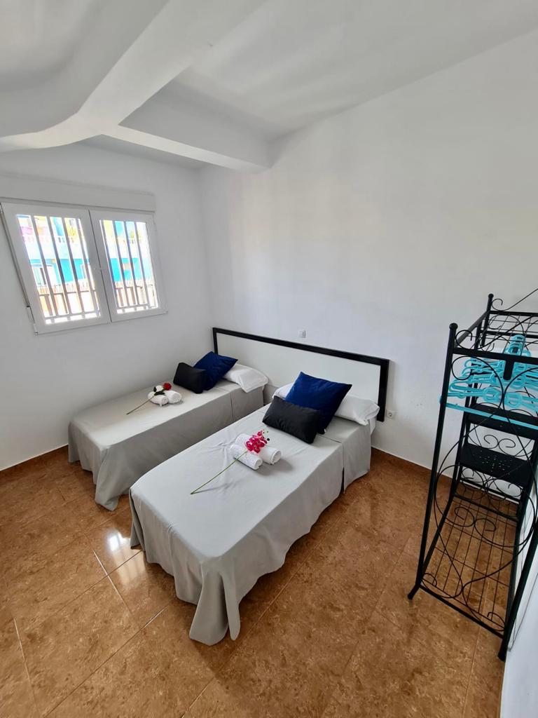 The bedroom features two individual beds covered with light-colored bedding, complemented by navy blue accent pillows. Natural light enters through two windows with subtle white blinds. A decorative metal shelf holds items in the corner, enhancing the room's functionality.