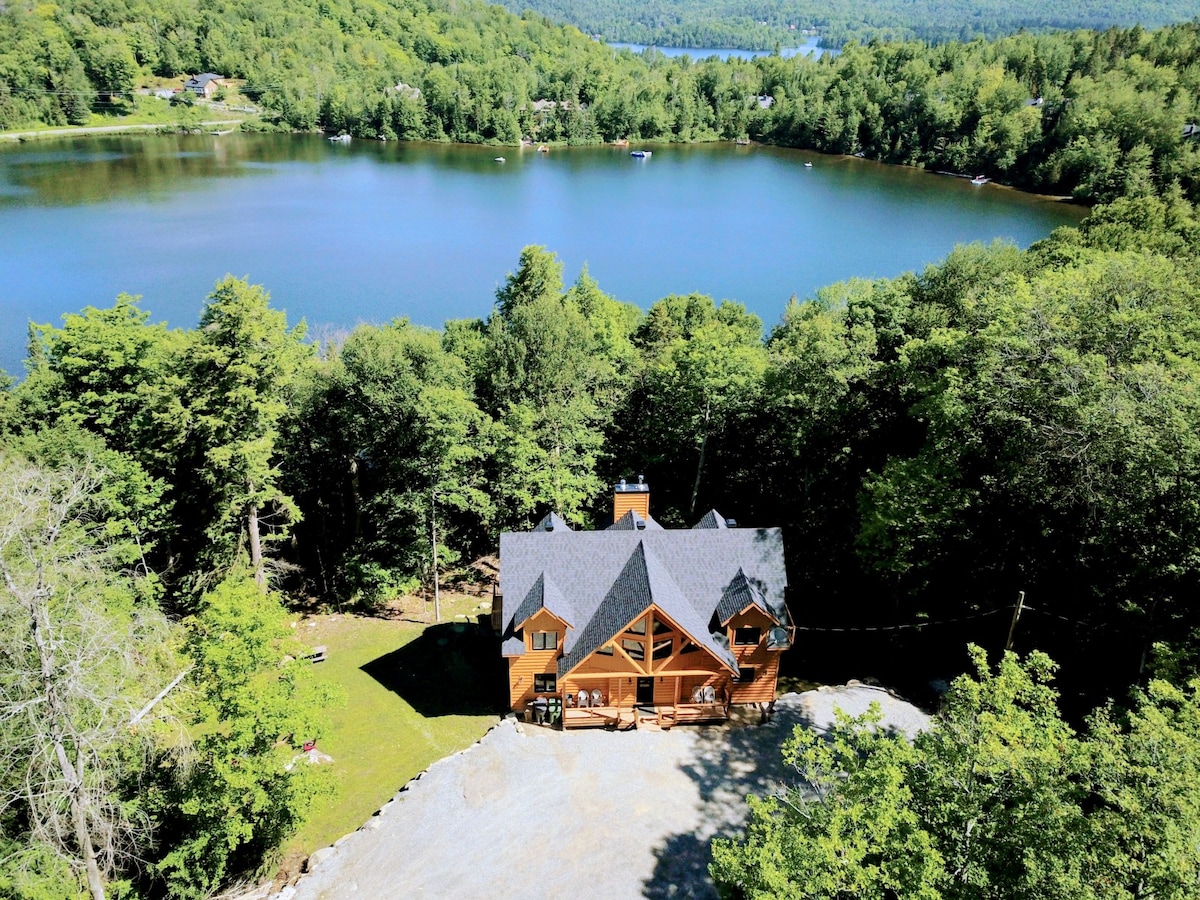 The chalet is positioned on a large wooded lakeside lot, surrounded by lush greenery. A clear lake is visible in the background, reflecting the vibrant landscape. The architecture features natural wood tones and an inviting roofline, creating a harmonious blend with the environment.