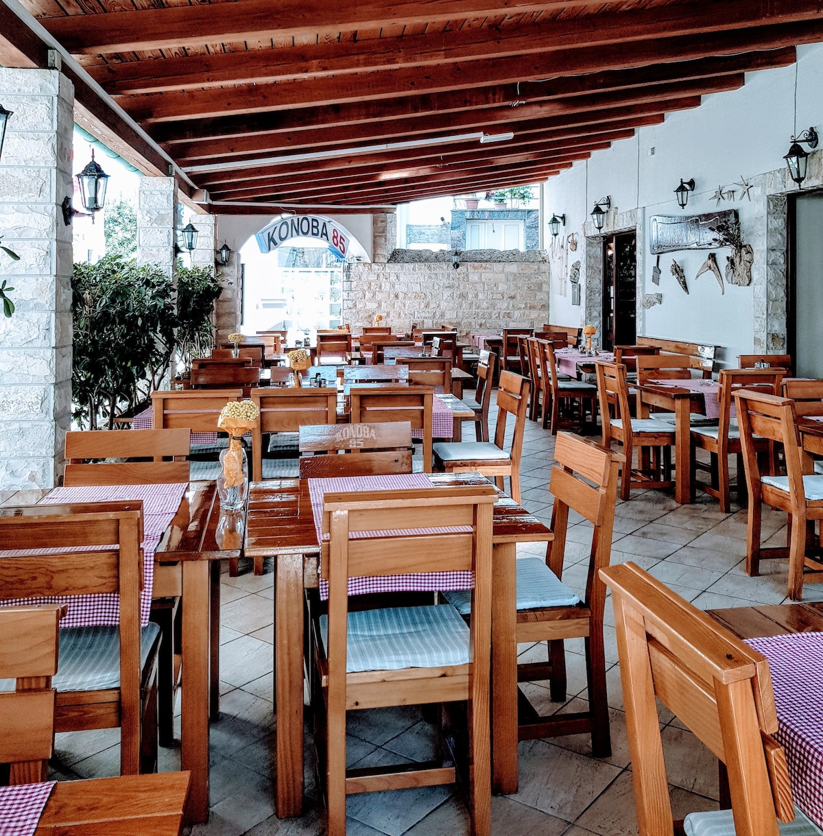 An outdoor dining area is depicted, featuring numerous wooden tables with matching chairs arranged under a wooden roof. Each table is set with checkered cloths and adorned with small flower vases. Soft natural light filters through the adjacent greenery, enhancing the relaxed ambiance.