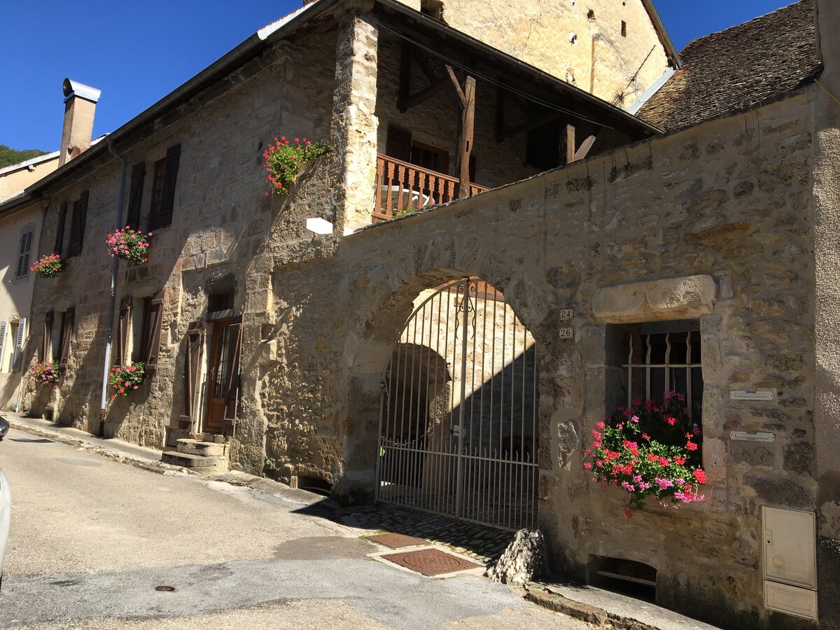 An aged stone building features a charming façade with flower boxes adorned by colorful blooms. An arched entrance provides access to a gated area. The rustic architecture reflects historical character, while wooden balconies add to the structure's unique design.