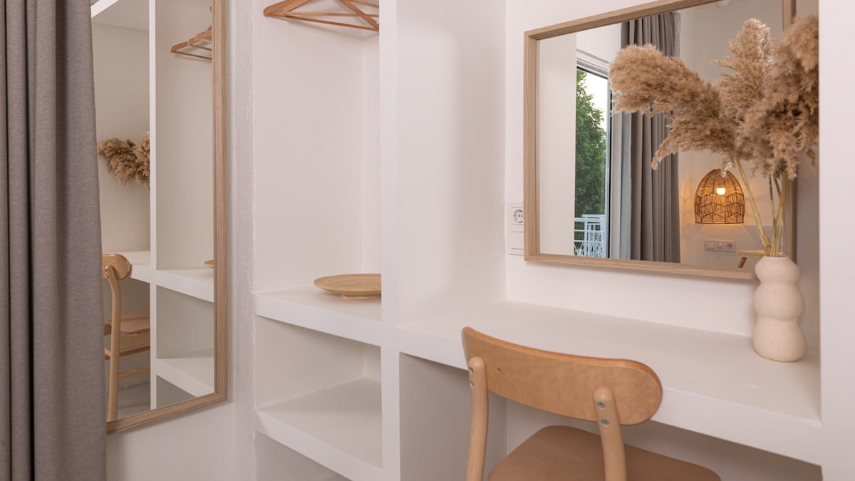 A well-lit vanity area is shown, featuring a smooth white tabletop with a wooden chair and a decorative vase holding dried flowers. Two mirrors reflect the space, while soft drapes frame the view. Natural light highlights the minimalist design.