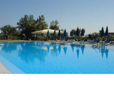 A large swimming pool glistens under a clear blue sky, surrounded by lounge chairs and vibrant blue umbrellas. Lush trees and greenery are visible in the background, adding a serene atmosphere to the outdoor space.