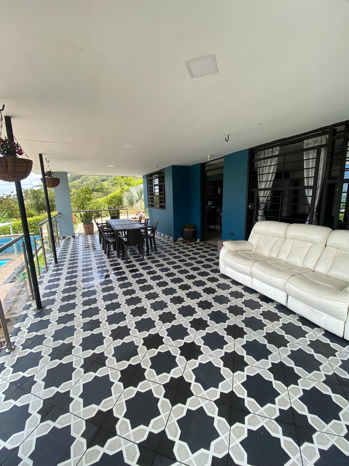 An outdoor patio space is visible, featuring a white sofa and a dining table set for six. The flooring showcases a patterned design, while large windows provide a view of the surrounding greenery. Hanging plants add a touch of nature to the area.