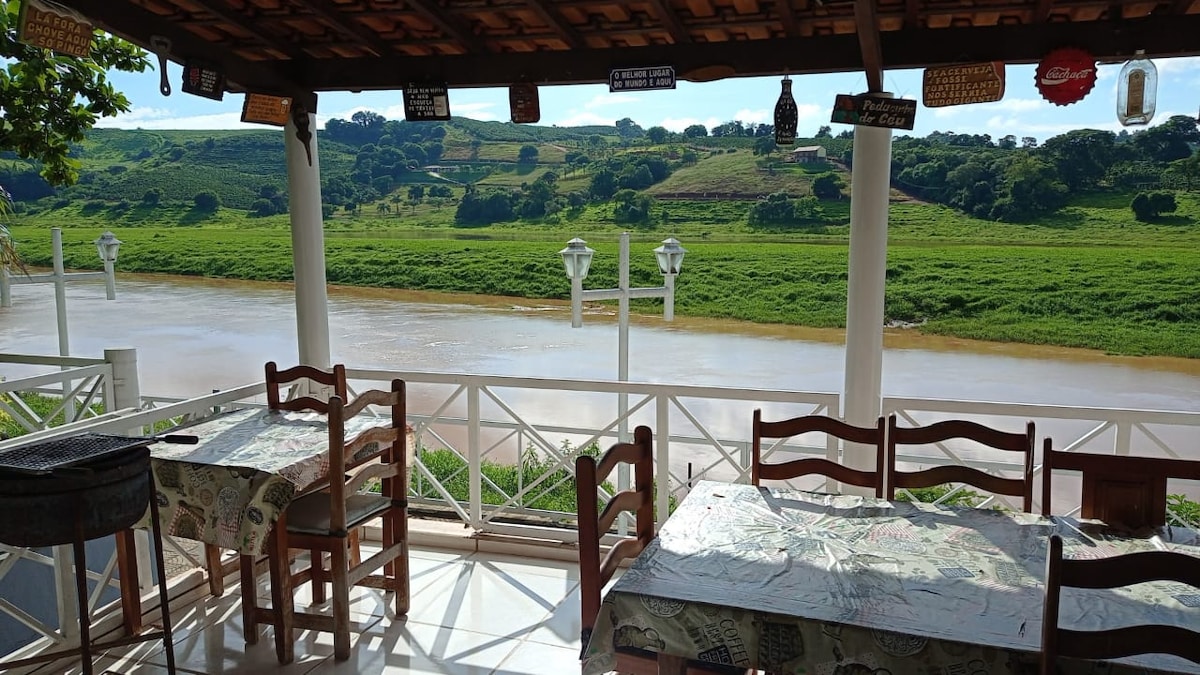 A covered terrace offers a serene view of a peaceful river, bordered by lush greenery. Wooden tables and chairs are set up for dining, with decorative signage hanging above. White railings frame the outdoor space, enhancing the connection to nature.