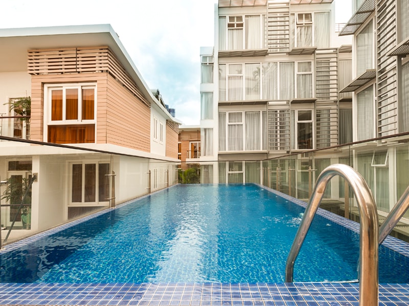 A clear, inviting swimming pool is framed by modern apartment buildings, featuring large windows that allow natural light to illuminate the space. The pool is surrounded by glass railings, enhancing the open atmosphere of the area.