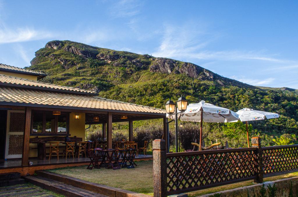 An outdoor seating area is available, featuring tables and umbrellas set against a backdrop of lush green hills. The well-maintained garden is complemented by a wooden fence, and a cozy patio is visible beside the main structure.