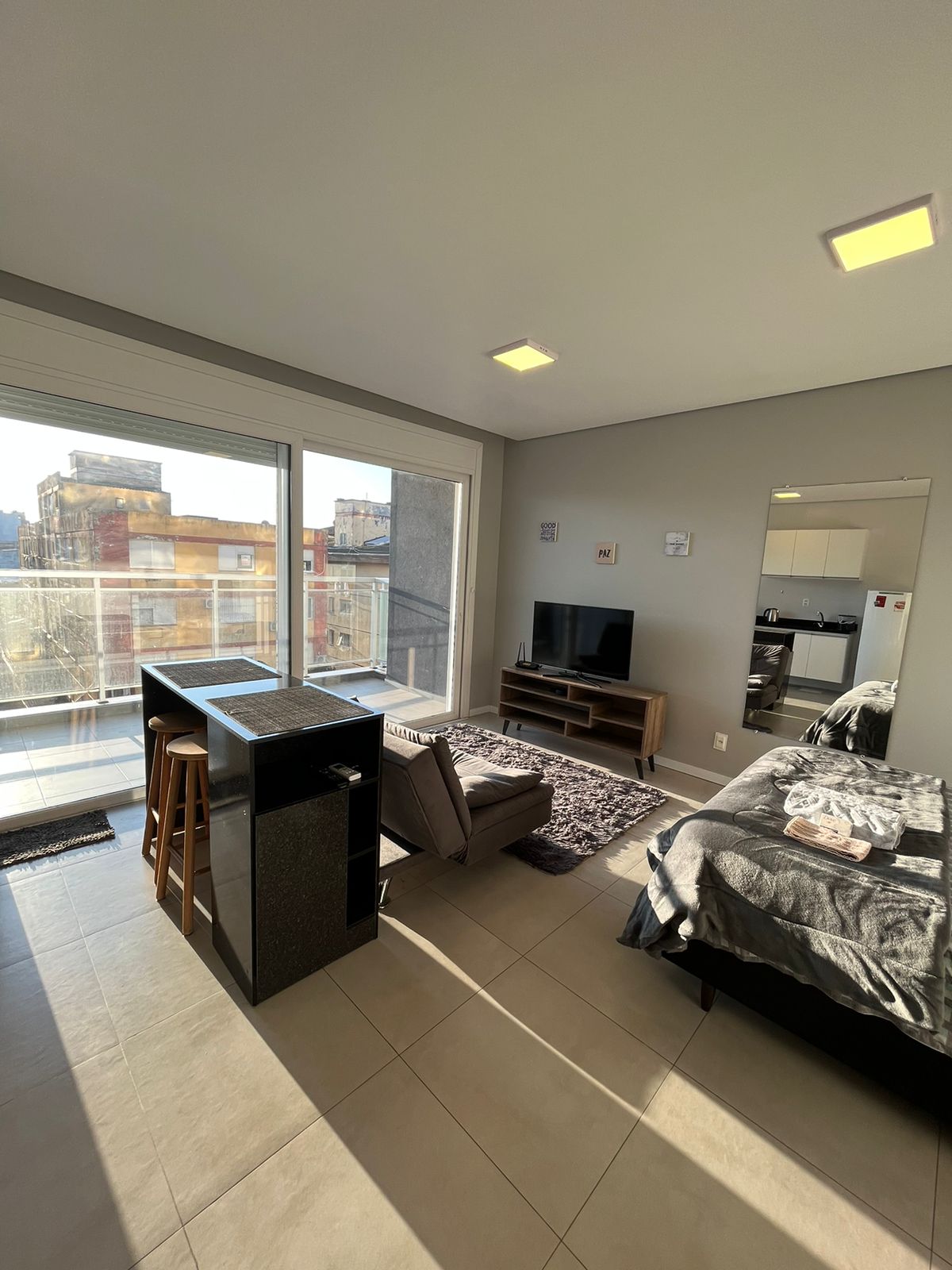 An open studio space is displayed, featuring a comfortable bed beside a modern entertainment unit with a television. A small dining area with two barstools is positioned near expansive glass doors, inviting natural light and offering views of the city beyond.