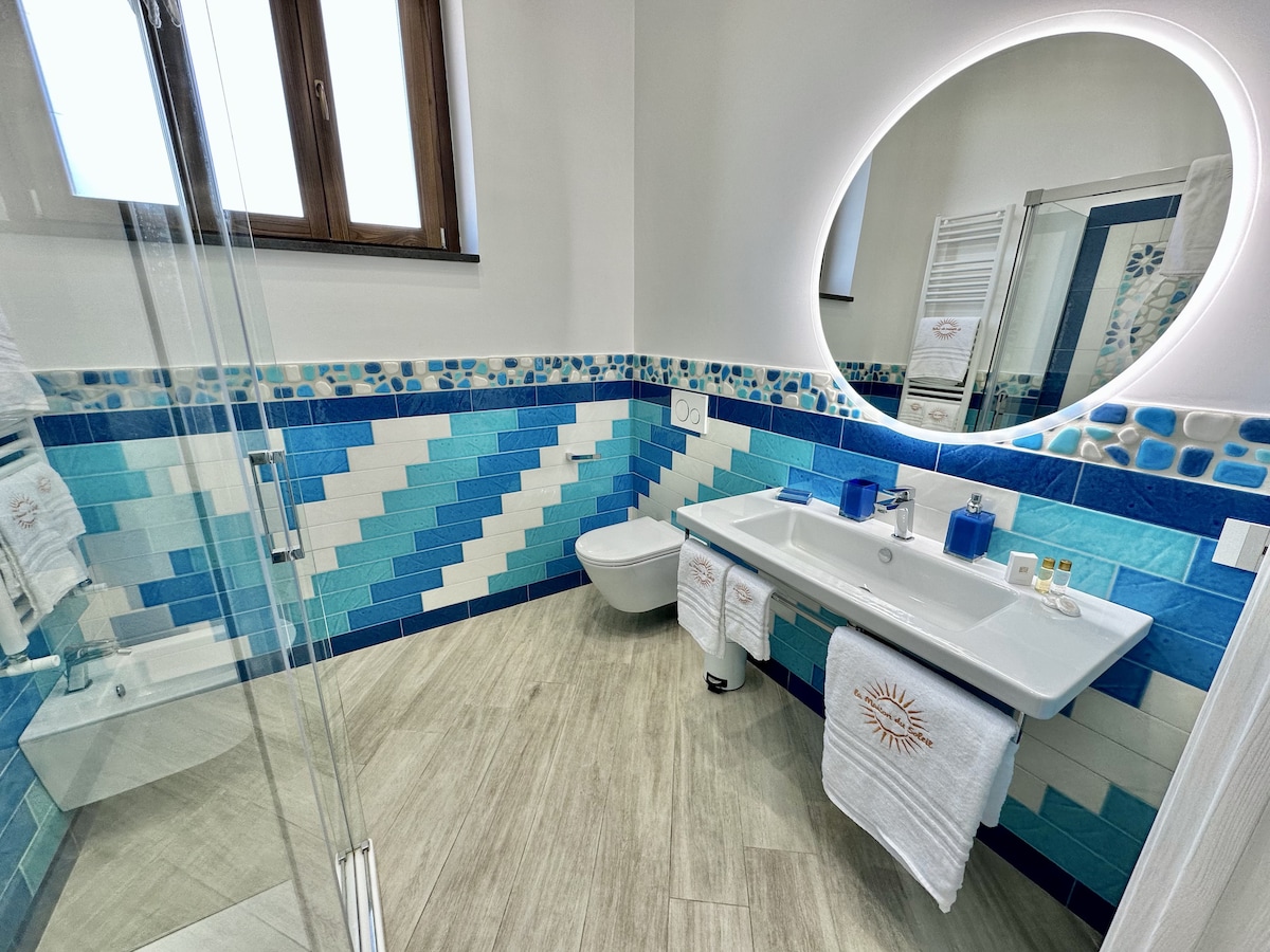 The bathroom features a spacious layout with a walk-in hydromassage shower and a modern sink area. A large mirror complements the colorful tiled walls, which showcase shades of blue and turquoise. Natural light filters through a window, enhancing the clean design.