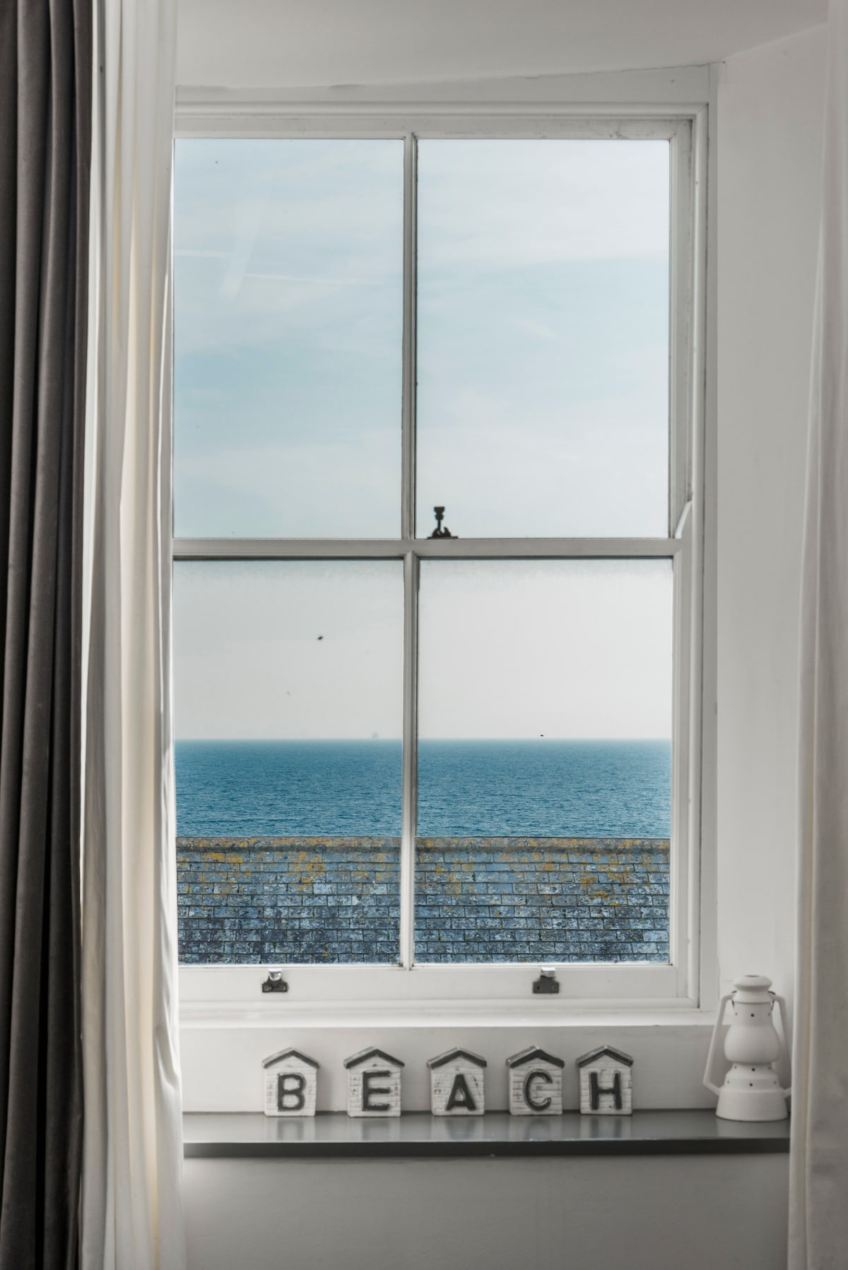 A large window frames a view of the calm sea, revealing soft blue waters merging with the horizon. The sill displays decorative letters spelling 'BEACH,' complemented by soft drapes in light and dark tones, enhancing the serene ambiance.