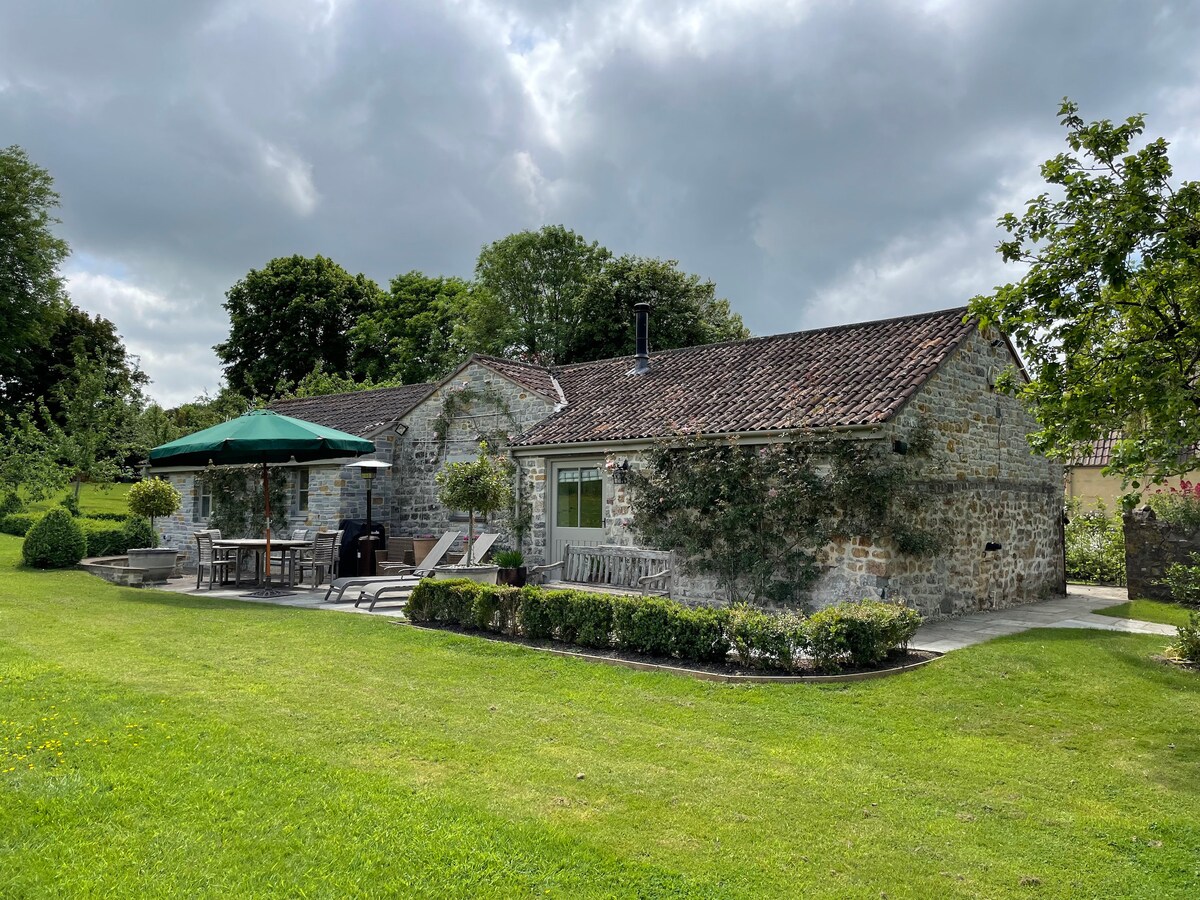 The exterior of a charming stone barn features a spacious terrace with a large umbrella and seating. Lush green grass surrounds the building, accompanied by neatly maintained gardens. The architecture reflects a blend of rustic charm and contemporary design, with ample windows allowing for natural light.