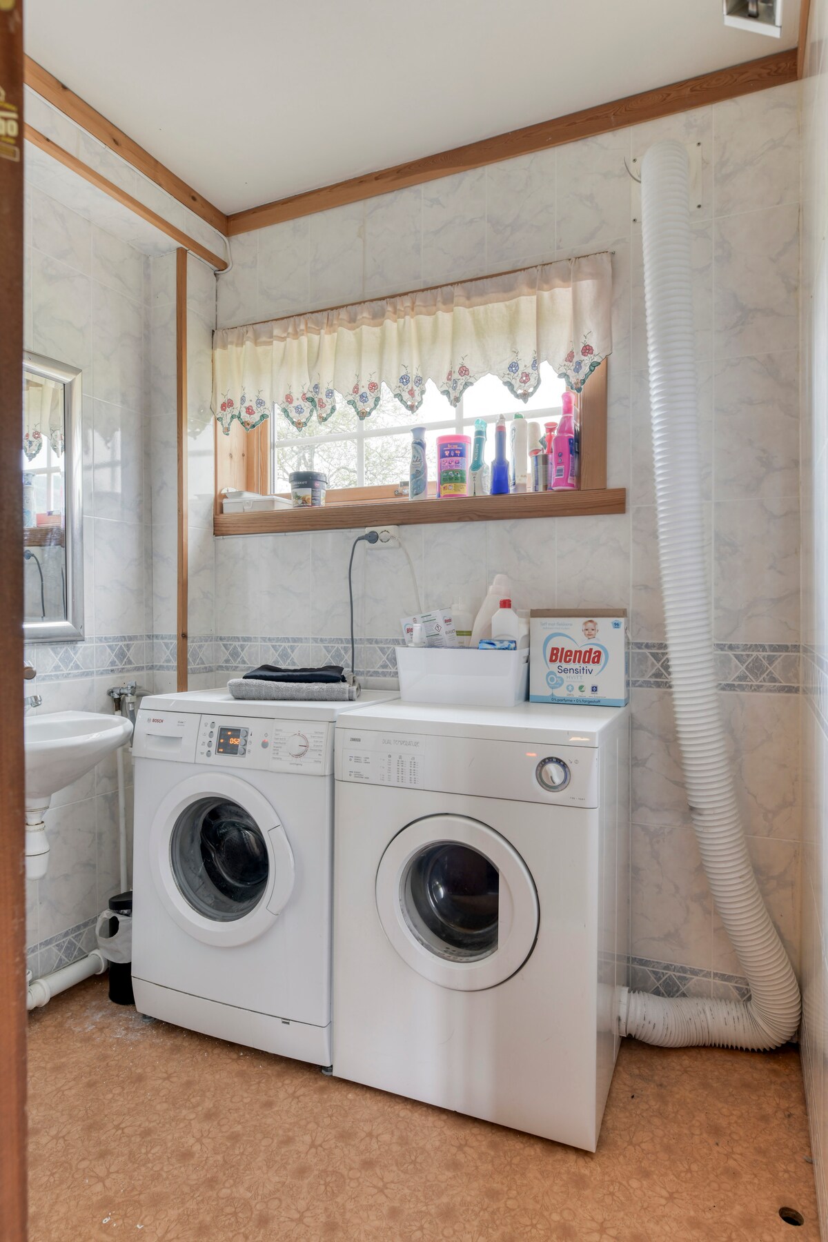 A laundry area features two white washing machines side by side, equipped with controls on the front. A small sink is positioned above a beige countertop, with cleaning supplies neatly arranged on top. Natural light filters in through a window adorned with lace curtains.