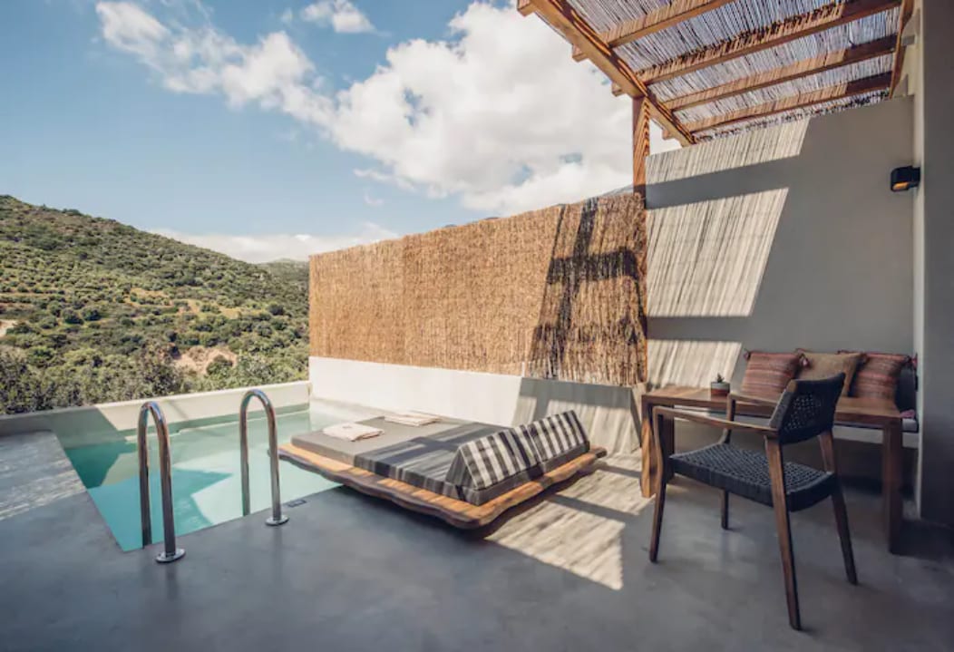 A sun-drenched terrace features a private swimming pool with a view of green hills. Outdoor seating includes a lounge mattress with a striped pattern and a black chair. Natural elements like wooden beams and a straw cover create a relaxing atmosphere.