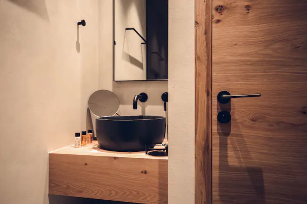 A bathroom features a natural wood vanity with a sleek black sink. A round mirror reflects the space while modern black fixtures are visible. A wooden door leads into the room, completing the minimalist aesthetic of the area.
