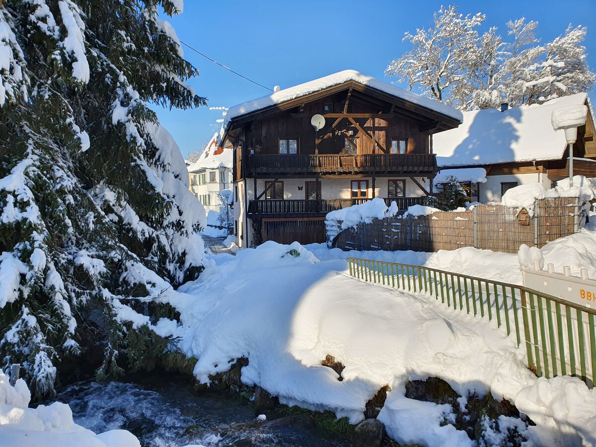 A charming wooden chalet is nestled amidst a snowy landscape, partially surrounded by tall evergreen trees. Snow blankets the ground and rooftops, while a gentle stream runs alongside the property. The clear blue sky above enhances the serene winter scenery.