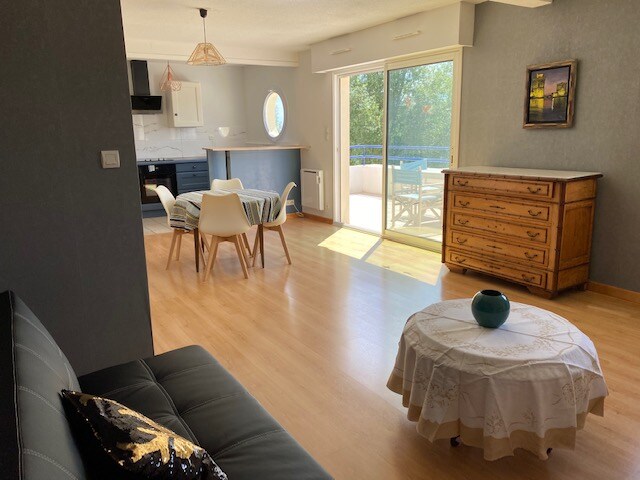 A spacious living area features a round dining table with four chairs and a decorative light fixture overhead. A comfortable sofa is positioned near a coffee table adorned with a small decorative piece. Large glass doors provide access to an outdoor terrace, allowing natural light to fill the room.