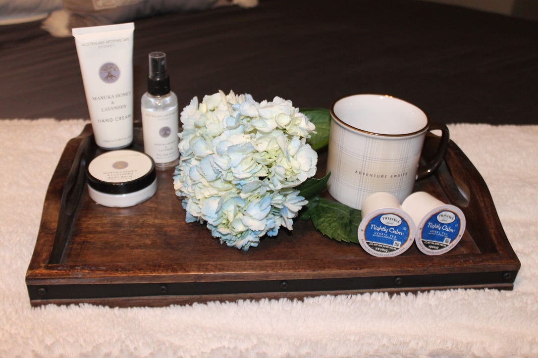 A wooden tray showcases a collection of personal care items, including hand cream, body spray, and two small containers. A white and blue floral arrangement is placed next to a ceramic mug on a textured white surface.