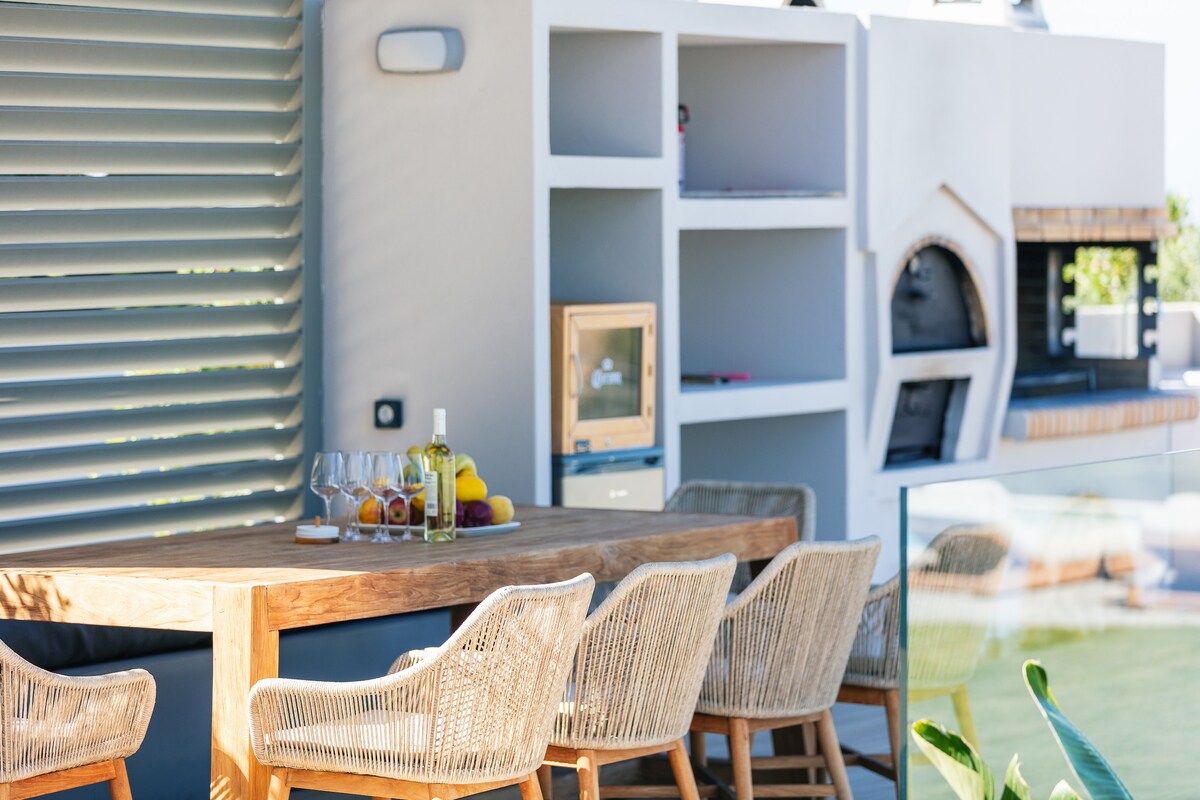 An outdoor dining area features a large wooden table surrounded by stylish wicker chairs. A barbecue station is visible in the background, complemented by built-in shelving. Fresh fruits and a bottle of olive oil are arranged on the table, enhancing the inviting atmosphere.