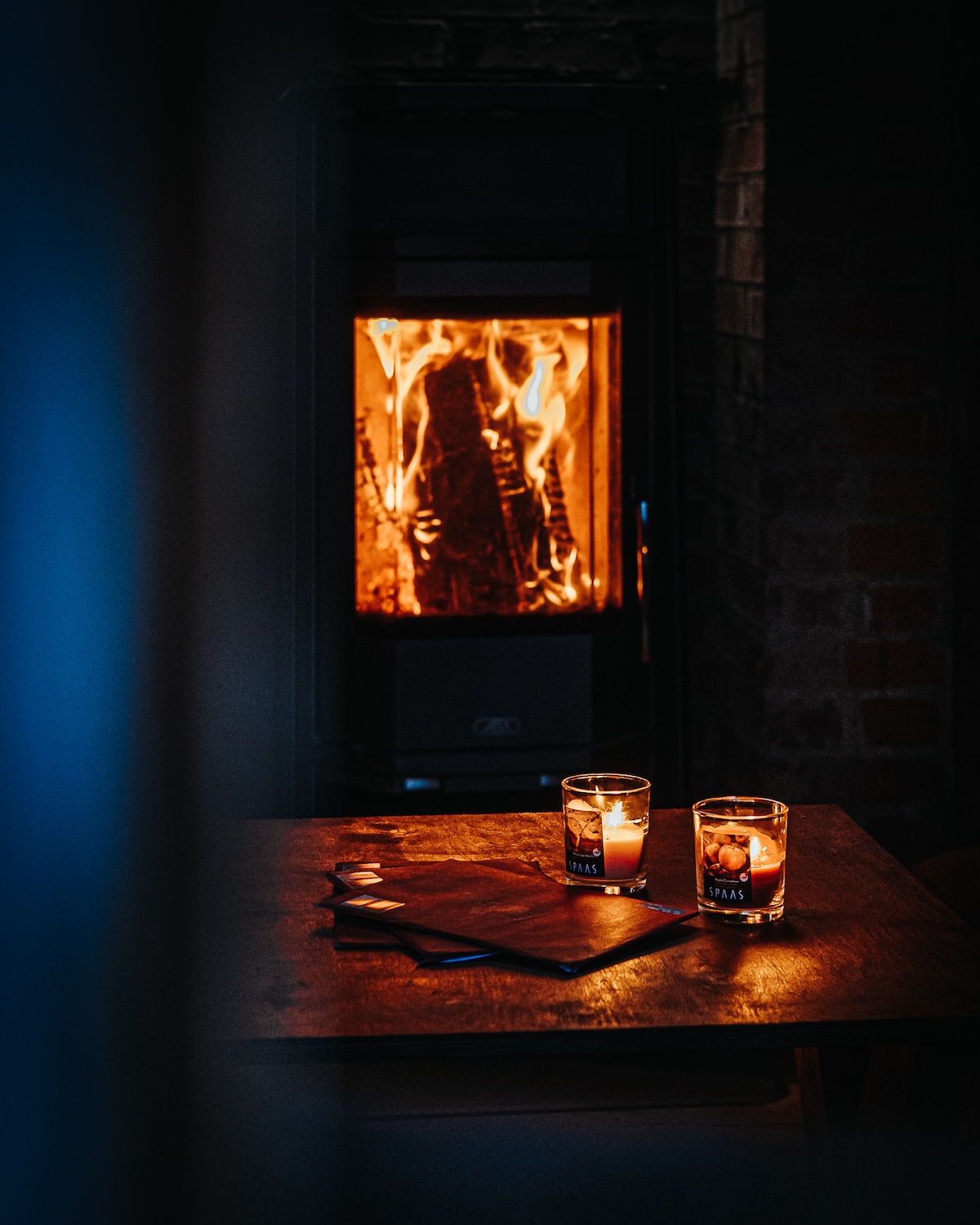 A cozy setting is created by a warm fireplace, where flickering flames are visible through a glass front. On a wooden table, two lit candles are placed beside a menu or notebook, providing a gentle illumination in the dimly lit room.