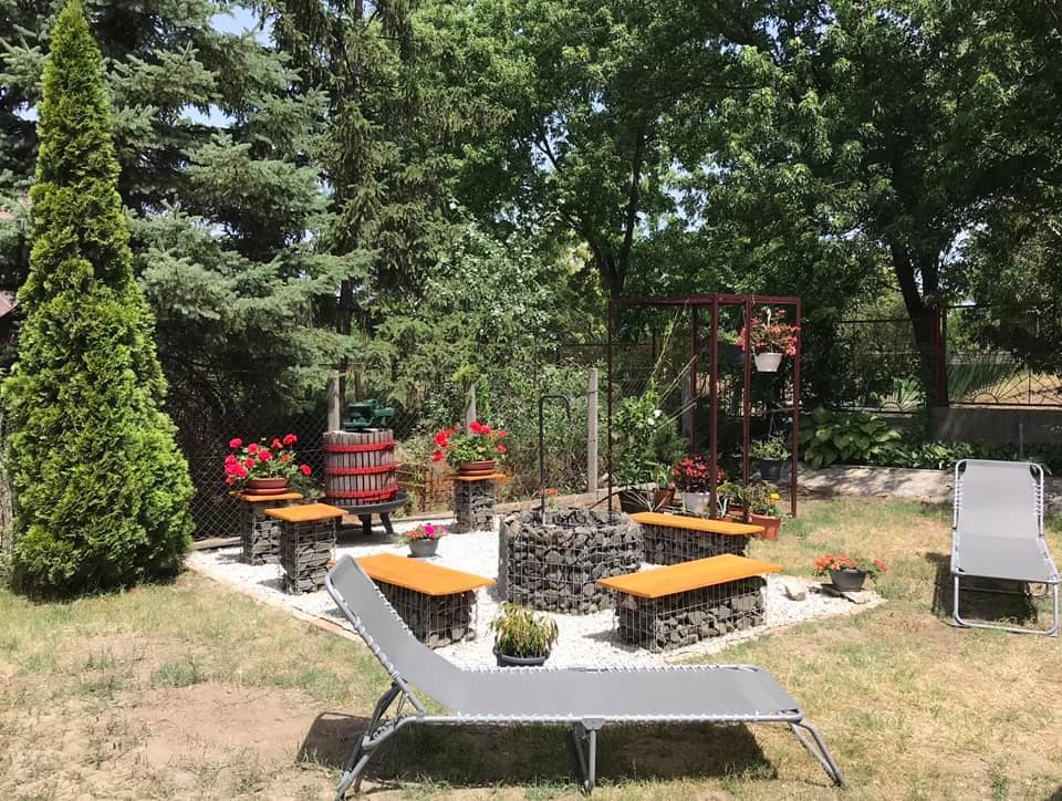 A serene garden area is highlighted, featuring a stone fire pit surrounded by wooden benches. Lush green trees provide shade, while planters with vibrant flowers enhance the landscape. A comfortable lounge chair is positioned nearby, inviting relaxation in the natural setting.