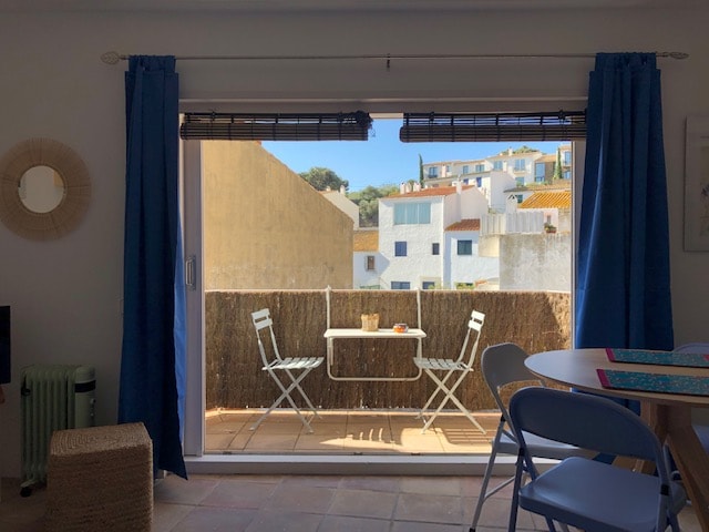 A serene view of a small balcony is captured, featuring a round table with two chairs. Sunlight filters through the open sliding door, illuminating the space. The backdrop includes neighboring buildings with terracotta roofs, while plant life adds a subtle charm to the scene.