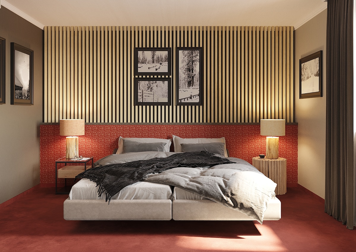 A spacious sleeping area features a large bed with a light-colored upholstered headboard. Two bedside tables with lamps provide soft lighting, while wall-mounted photographs of winter landscapes add a decorative element. Warm tones are enhanced by a red accent wall and plush carpet.