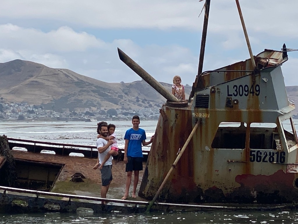 Three individuals are observed on the remains of a rusted shipwreck, with mountainous terrain visible in the background. One person is standing on the ship's bow while another holds a child. The calm water reflects the ship's weathered texture, creating a serene coastal atmosphere.