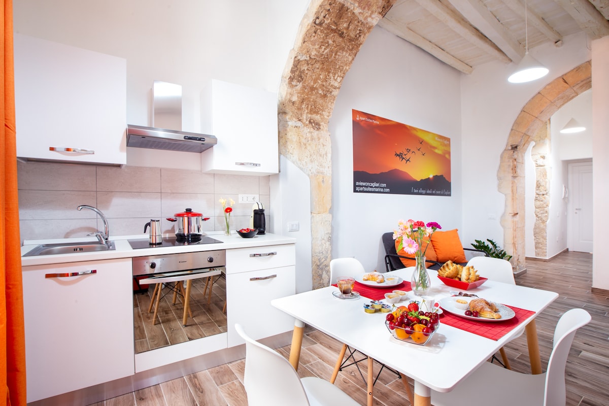 A well-equipped kitchen area features modern white cabinetry and stainless steel appliances. A dining table is set with a colorful display of fresh fruit and pastries. Arched stone walls add character, while natural light brightens the space through a nearby window.