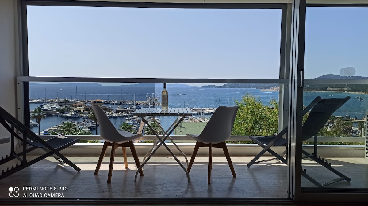 A terrace with a clear view of the sea and marina is showcased. Two white chairs and a small table are positioned to take advantage of the scenic surroundings. Natural light fills the space, highlighting the outdoor area while the calming blue of the water is visible in the background.