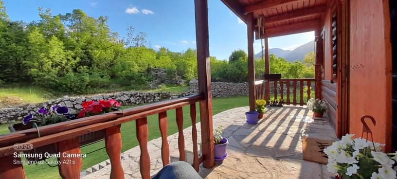 A serene porch is framed by wooden railings, leading to a well-maintained garden. Colorful pots with flowers add a vibrant touch, while the stone floor provides a natural feel. Lush greenery and distant hills create a peaceful backdrop, ensuring a tranquil outdoor experience.