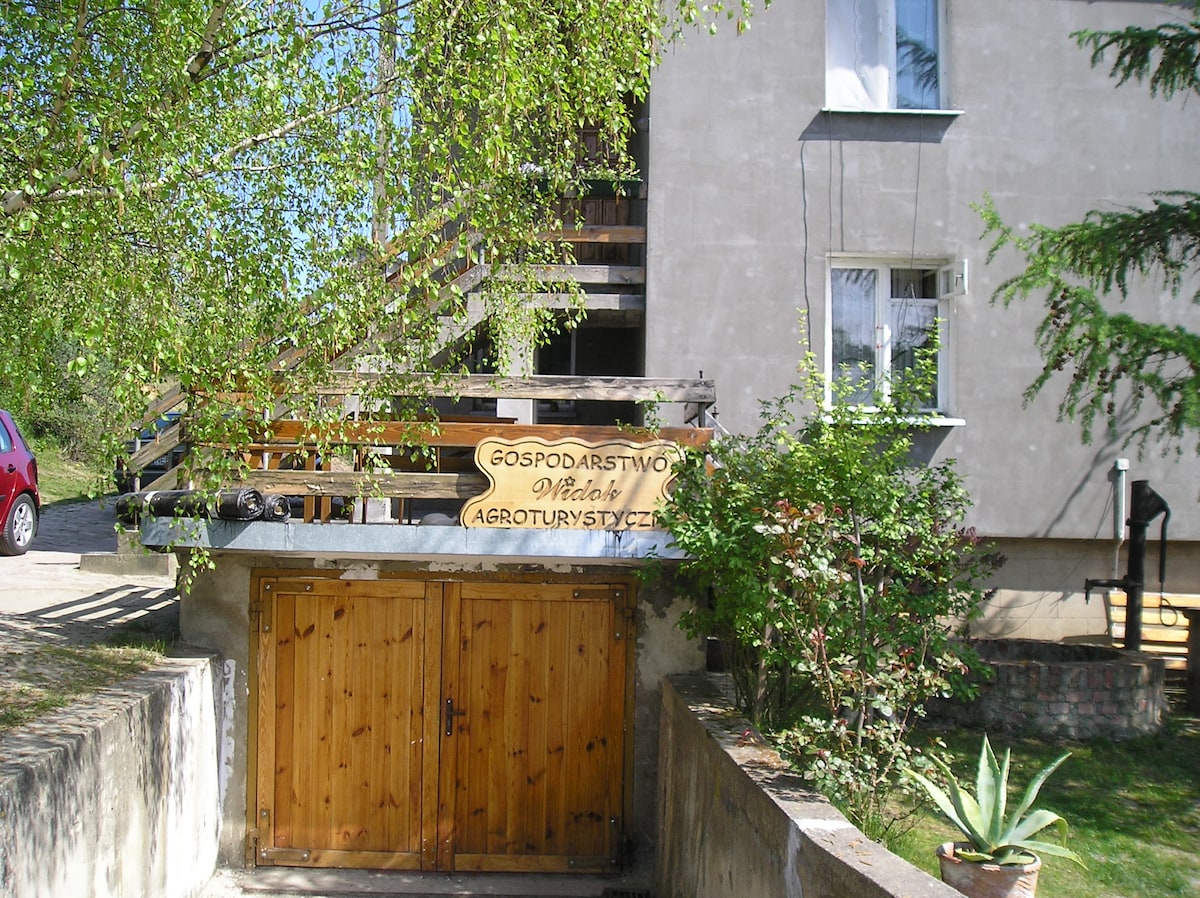 The entrance to the property features a wooden sign that reads 'Gospodarstwo Agroturystyczne.' A parked car is visible nearby, while green foliage frames the area. The building's exterior is complemented by a staircase leading to a terrace above.