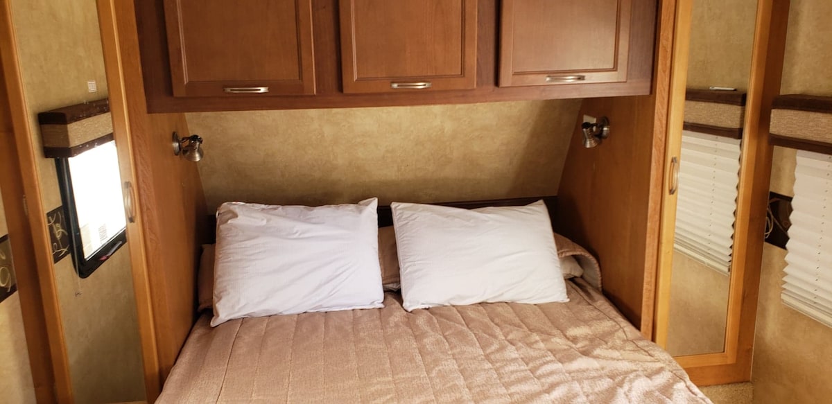 A cozy sleeping area features a spacious bed with a neutral-colored quilt and two white pillows arranged neatly on top. Soft lighting is provided by bedside sconces, and cabinets above offer additional storage. The walls are finished in warm tones, enhancing the comfort of the space.