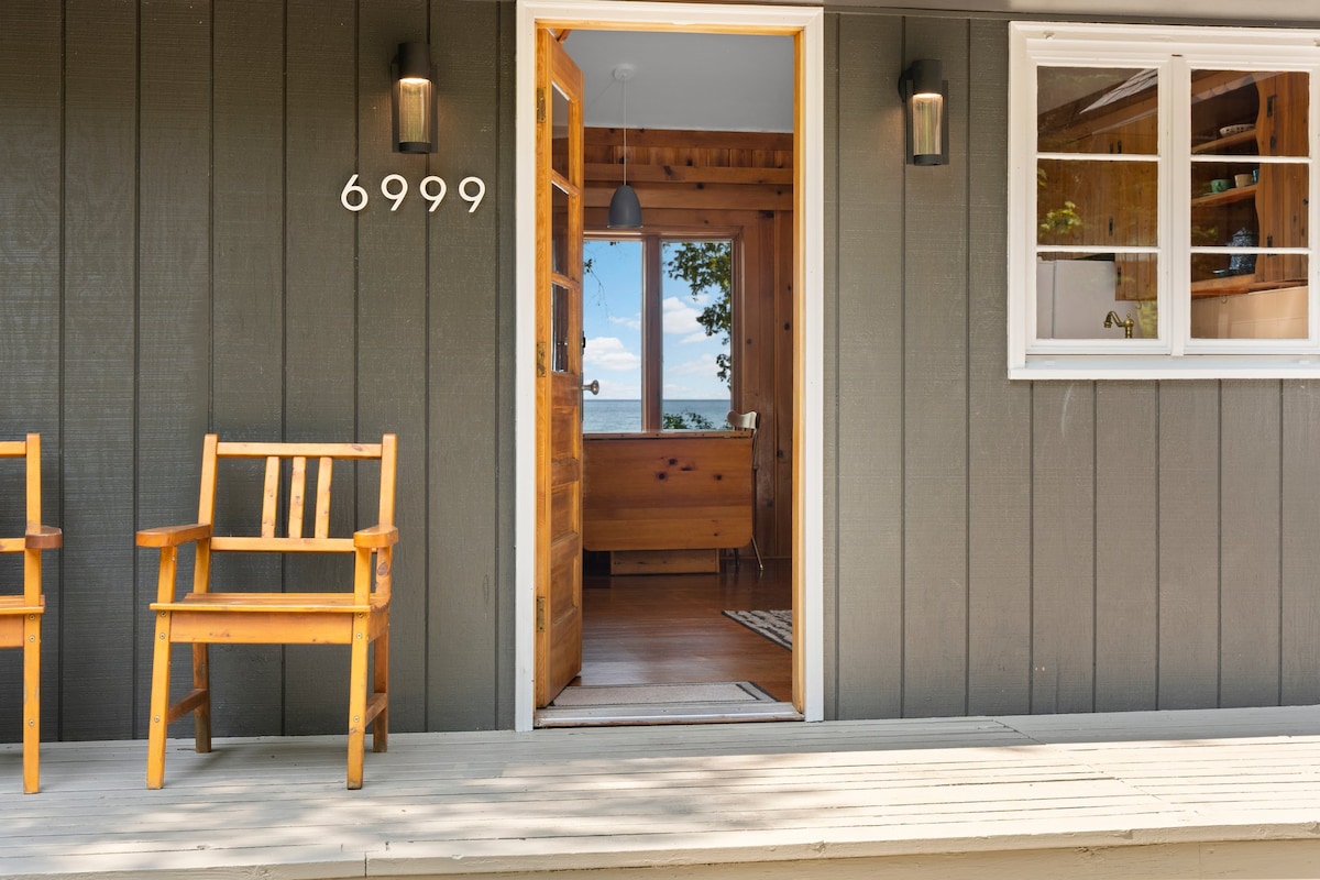 The exterior entrance of a cozy cottage is framed by two wooden chairs on a spacious porch. The inviting front door leads into a warm interior that showcases knotty pine accents. Natural light spills through the window, revealing a glimpse of the lake beyond.