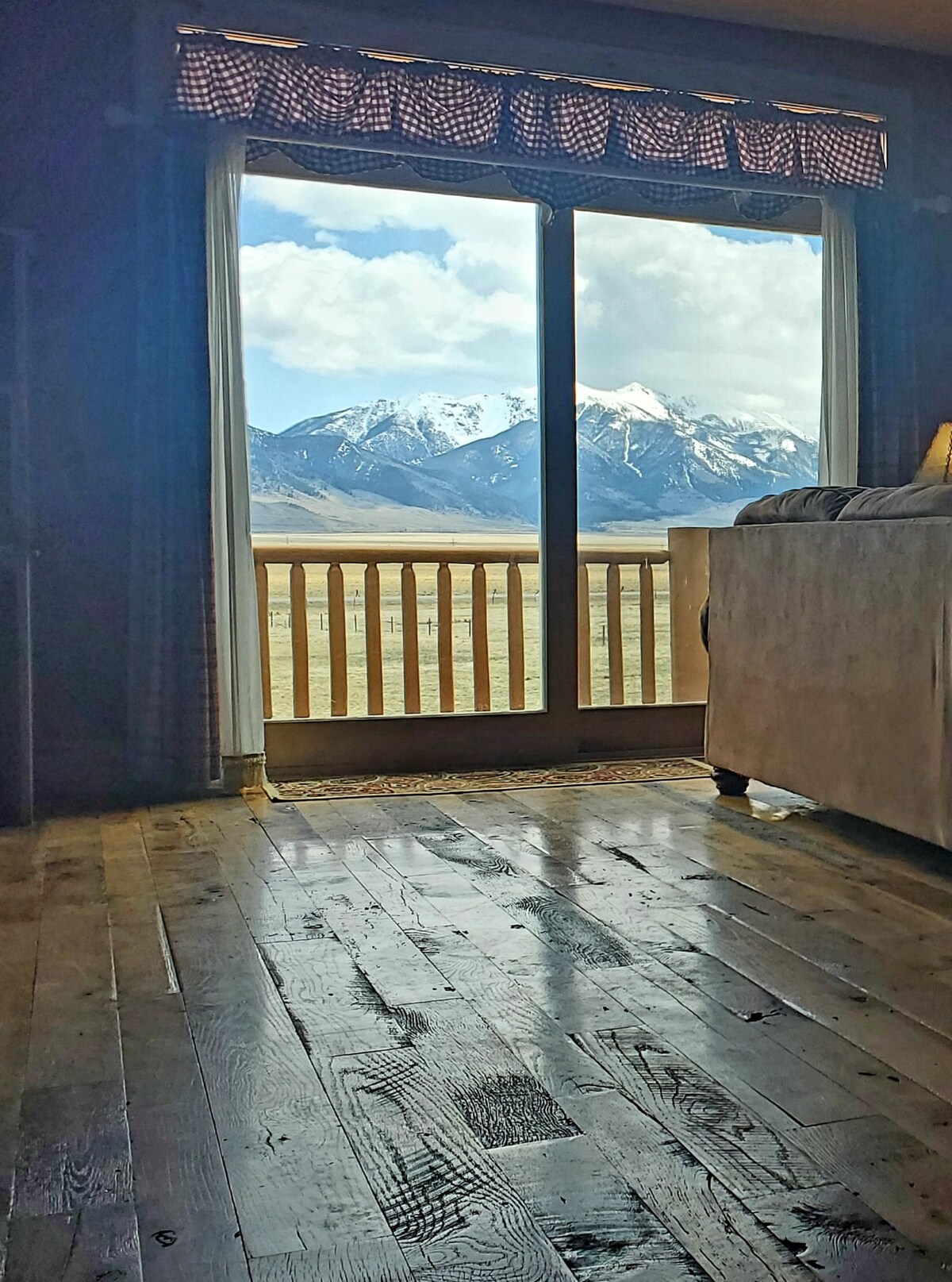 A wide glass door provides a view of snow-capped mountains framed by white clouds. Natural light floods the room, highlighting the polished wooden floor. The balcony railing is visible, enhancing the connection to the picturesque landscape beyond.