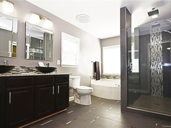 A modern bathroom features dark cabinetry and dual sinks with bowl-style basins. A glass-enclosed shower with a textured tile accent wall is visible alongside a white, sunken bathtub. Natural light is let in through a nearby window, enhancing the spacious feel.