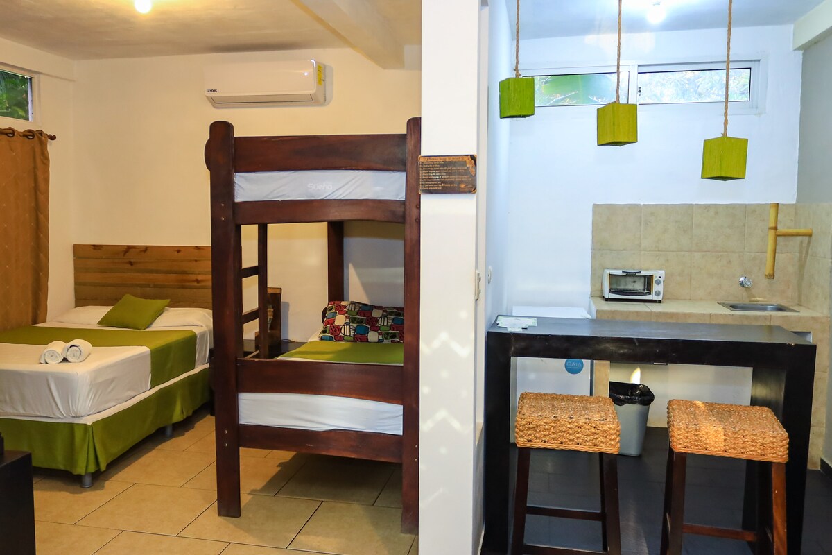 A well-appointed room features a spacious bed alongside a wooden bunk bed. The kitchenette area includes modern appliances and two seated stools. Natural light filters through the window, enhancing the neutral tones and simple decor.