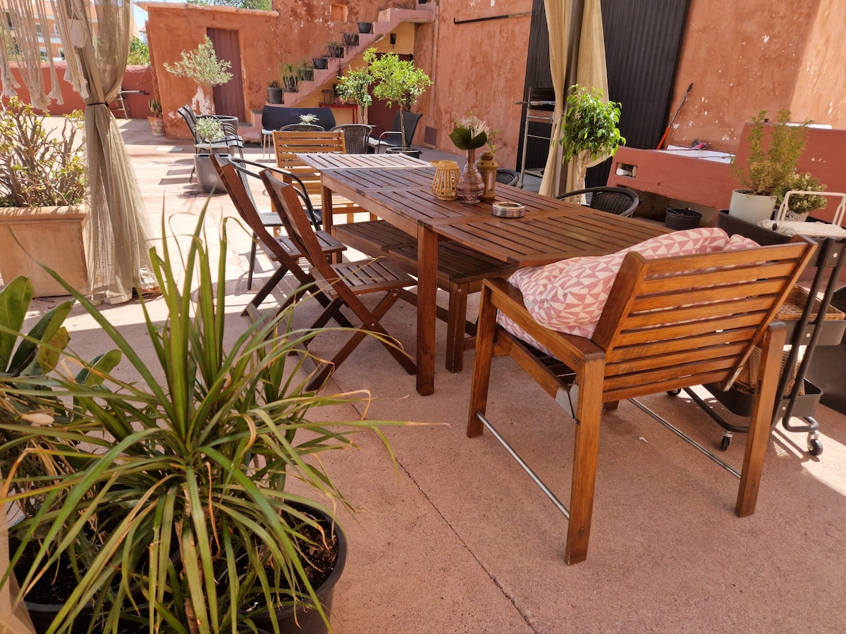 An outdoor terrace is depicted with a wooden dining table surrounded by several chairs. Potted plants are positioned around the area, and a staircase can be seen in the background, leading to an upper level. Sunlight brightens the space, enhancing its inviting atmosphere.