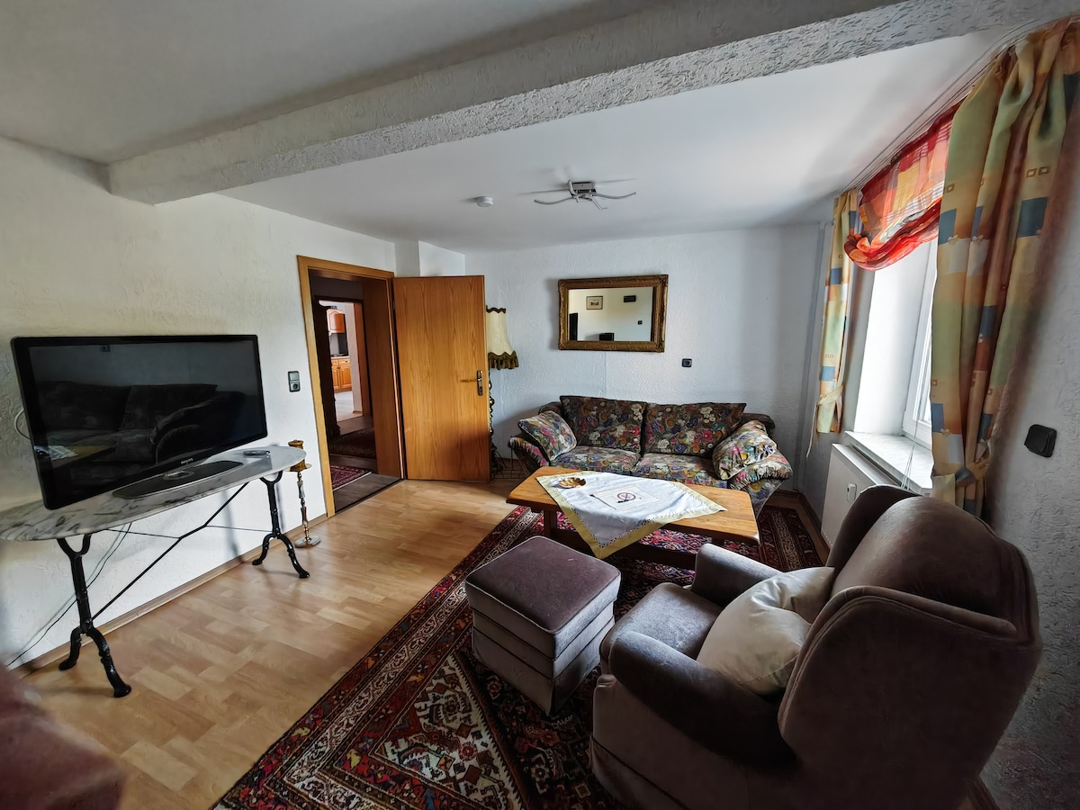 The living area features a comfortable seating arrangement, including a patterned sofa and armchair. A wooden coffee table is centered on a decorative rug, while a flat-screen television is positioned on a marble console. Colorful curtains frame the nearby window, allowing natural light into the space.