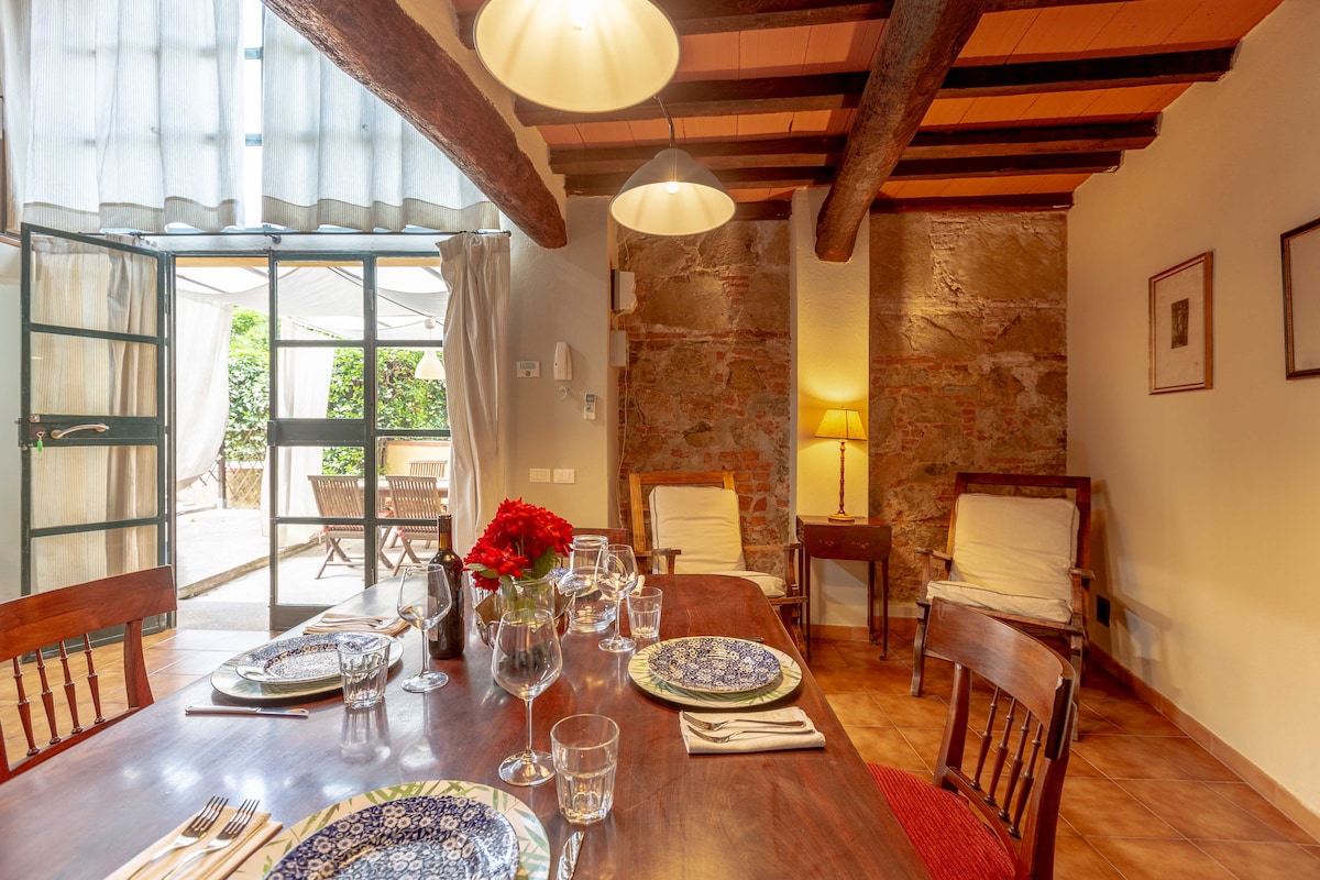 A dining area showcases a wooden table set for a meal, accentuated by decorative plates and glassware. Ambient lighting is provided by hanging lamps, while the rustic stone walls and exposed wooden beams create a warm atmosphere. A doorway leads to an outdoor space.