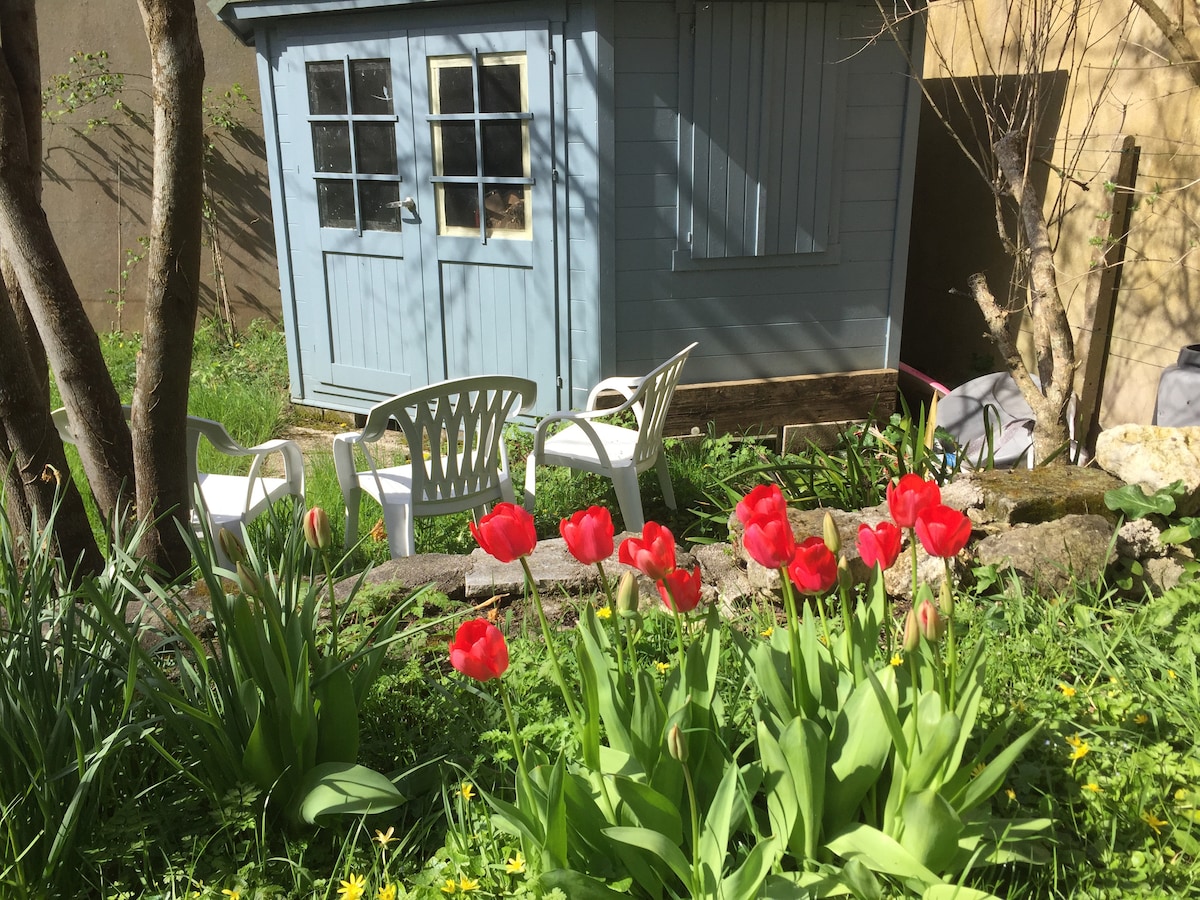 A quaint garden shed painted in a soft blue hue stands amidst vibrant red tulips and lush greenery. White plastic chairs are positioned nearby, offering a space to relax. The natural surroundings, including a mix of flowers and grass, create a peaceful outdoor atmosphere.