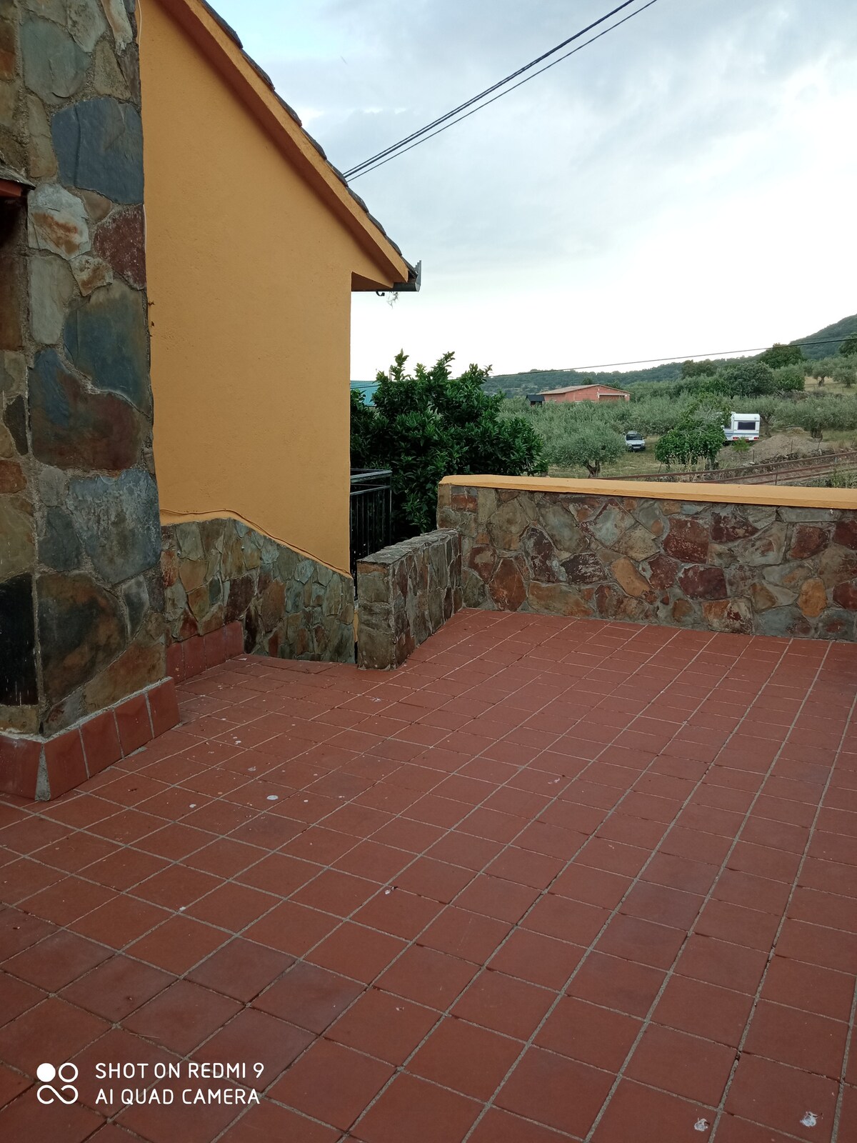 A stone-paved terrace area is presented, featuring warm-toned tiles and a low wall framed by greenery. The view beyond extends to rolling hills and open spaces, hinting at the natural surroundings. The structure of the building is visible in the background.