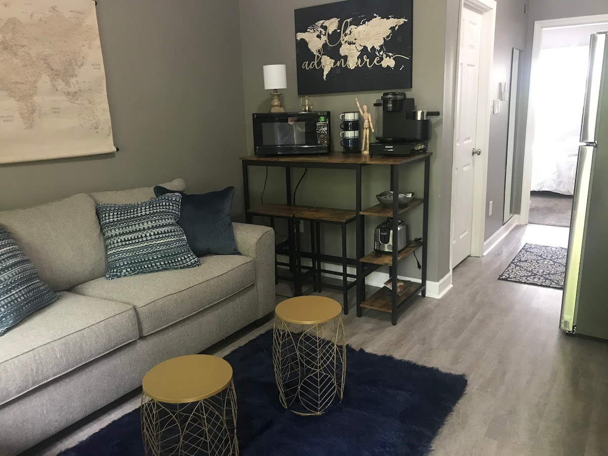 A comfortable living area is furnished with a light gray sofa adorned with decorative pillows. Two golden side tables sit on a soft blue rug, while a microwave and coffee station are organized on a wooden shelf against the wall. A map decorates the wall above.