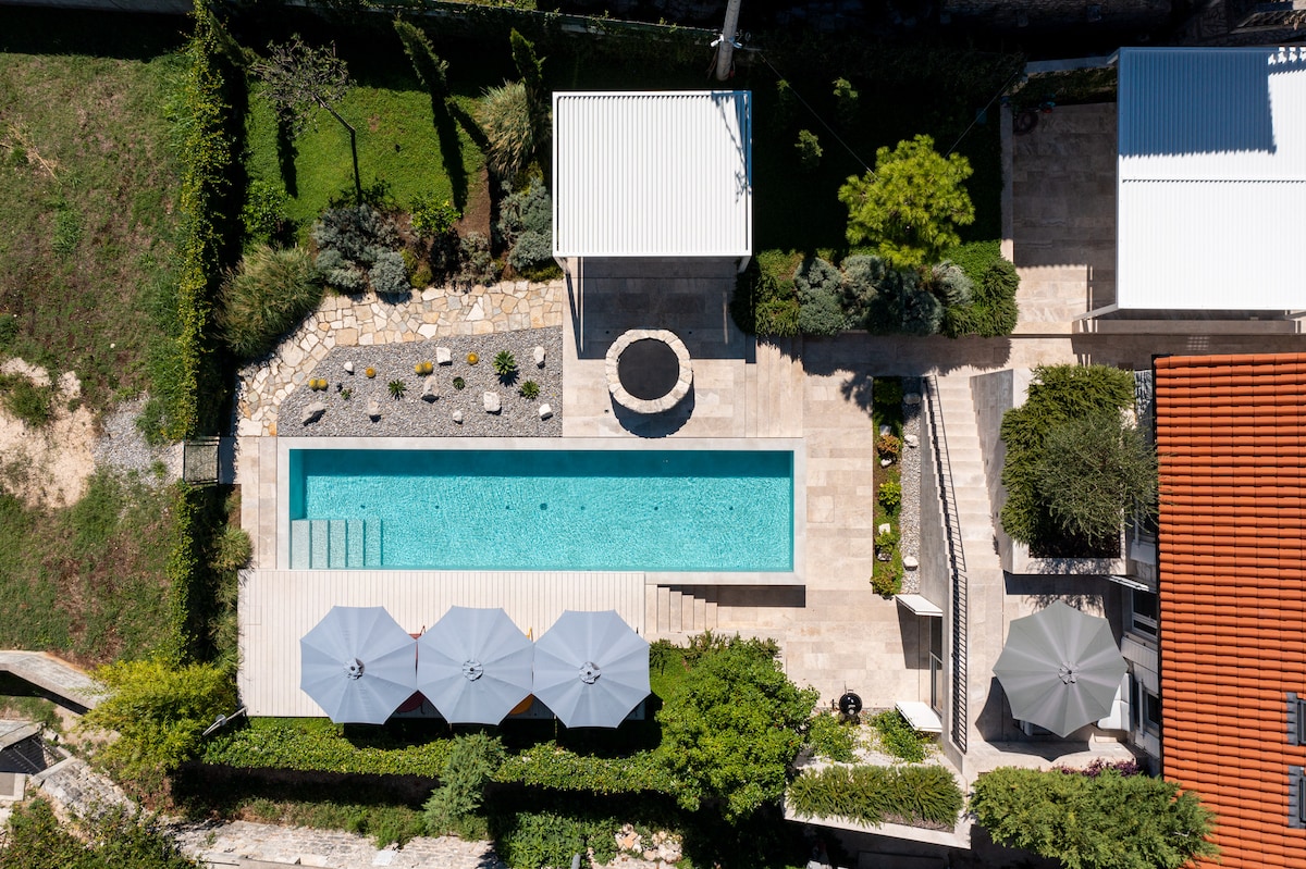 An aerial view captures a sleek swimming pool surrounded by sun loungers and umbrellas. A garden pathway leads to the pool area, bordered by greenery. Various outdoor seating is arranged nearby, offering multiple spaces for relaxation. The villa's modern design is evident throughout the property.