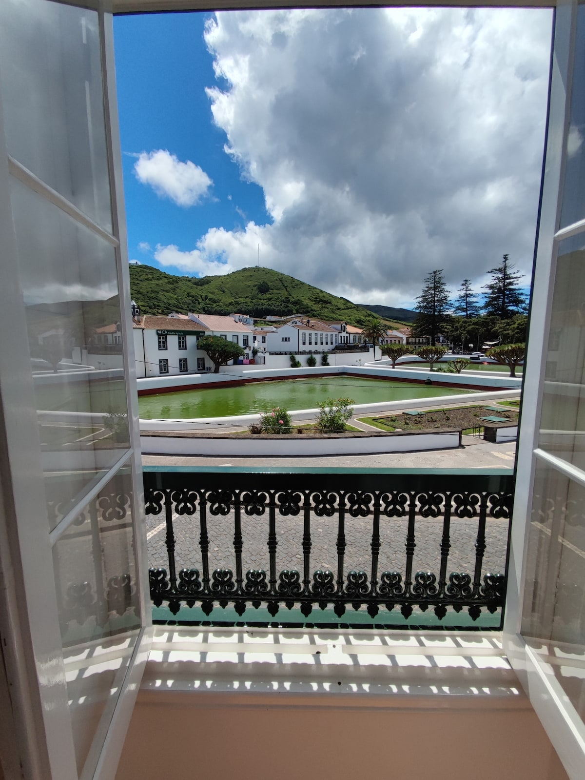 A view from an open window showcases a scenic landscape with rolling hills and a well-maintained garden. The decorative balcony railing adds character to the image, while the sunlight casts shadows on the floor, enhancing the tranquil ambiance.