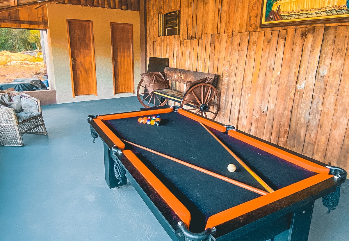 A billiards table occupies the center of a spacious area, surrounded by warm wooden walls. A vintage cart is positioned nearby, complementing the rustic decor. A soft seating arrangement is visible in the background, creating a relaxed atmosphere for leisure activities.