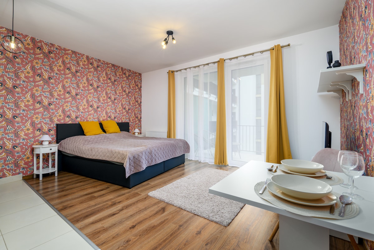 A spacious studio flat is depicted, featuring a king-size bed adorned with yellow accent cushions. A dining table is set for two, while large windows with flowing curtains allow natural light to illuminate the vibrant wallpaper. A soft area rug enhances the floor's warmth.