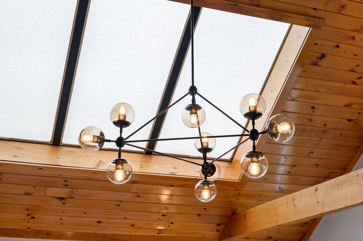 A modern industrial chandelier, featuring multiple glass globes, hangs from a wooden ceiling paneled with natural oak. The unique design adds a contemporary touch, while the glass provides ample illumination to the space. A skylight above enhances the natural light entering the room.