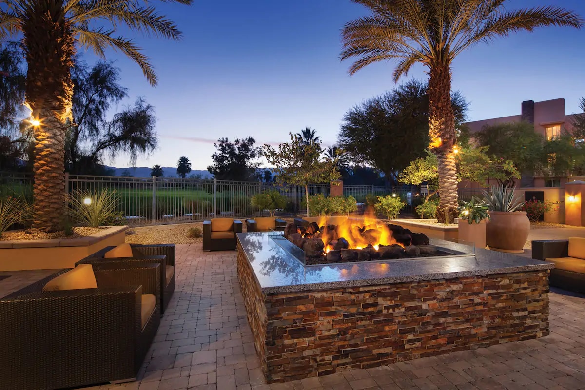 An outdoor seating area features several dark wicker chairs arranged around a stone fire pit, which is lit and providing warm illumination. Palm trees and well-maintained landscaping surround the space, with a green expanse visible in the background.