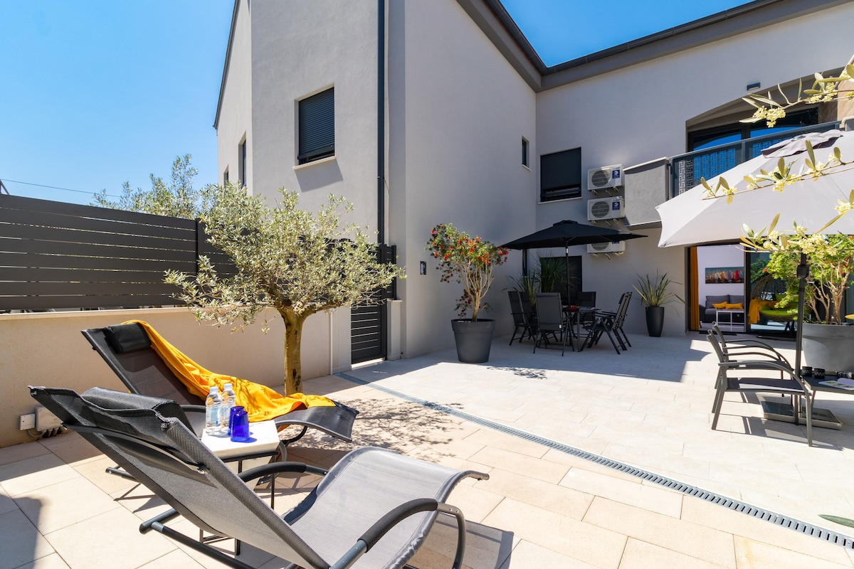 An outdoor patio area features comfortable lounge chairs and umbrellas, providing shade over a dining set. Potted plants add greenery to the space. The backdrop includes a modern building and a clear blue sky, creating an inviting and relaxing environment.